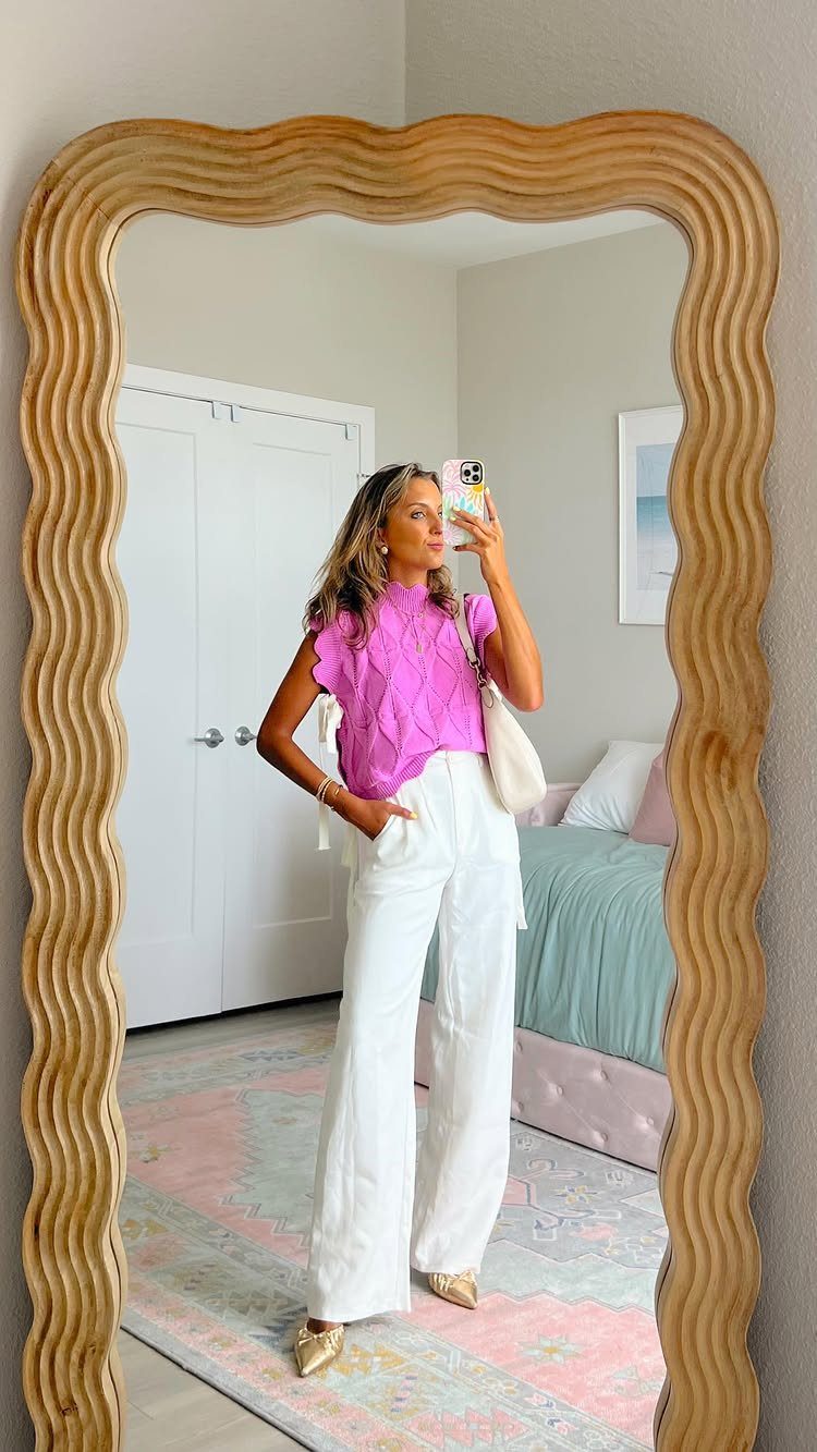 Woman in pink ruffled blouse and white wide-leg trousers posing in a wavy mirror.