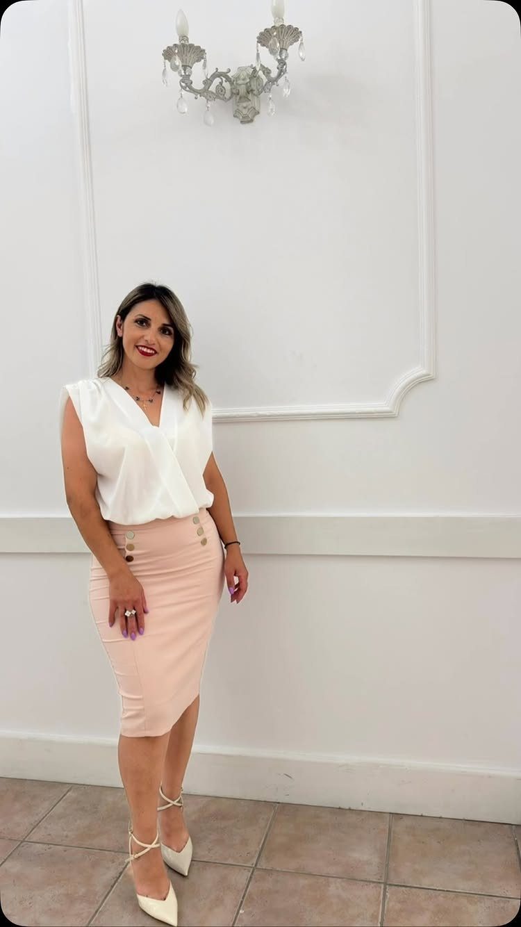 Woman in a white sleeveless blouse and peach pencil skirt posing against a white wall.