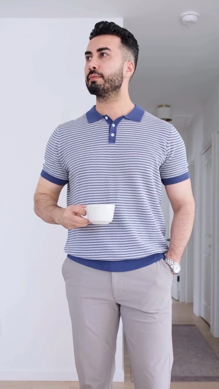 Man in a striped polo shirt and grey trousers holding a white cup indoors.