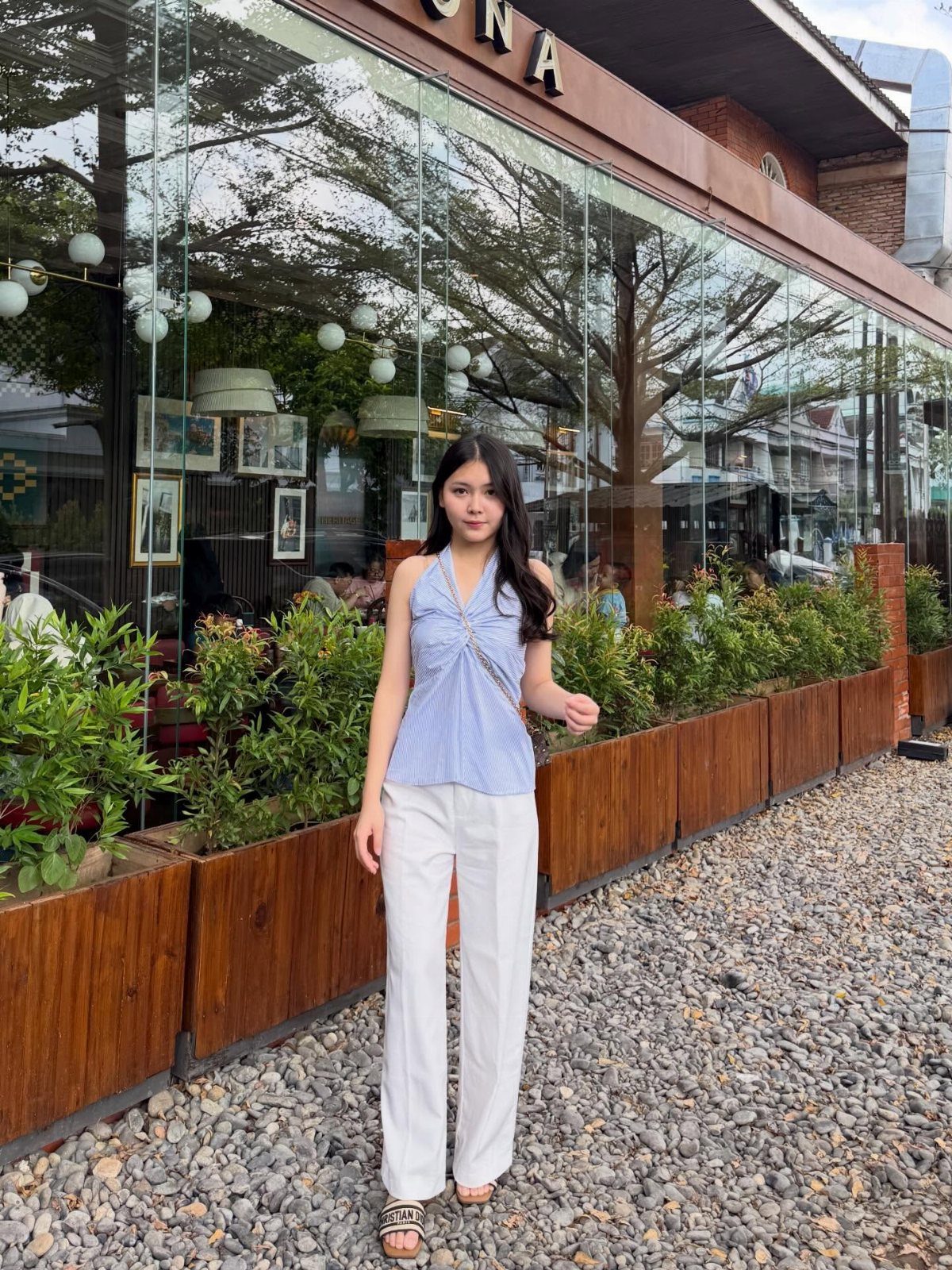 A young woman posing in a blue halter top and white trousers outdoors.