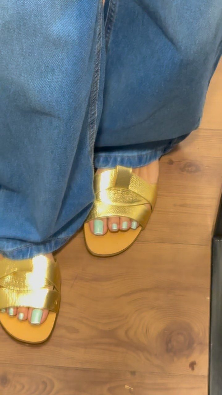 Gold metallic leather slide sandals worn with blue denim jeans on a wooden floor.