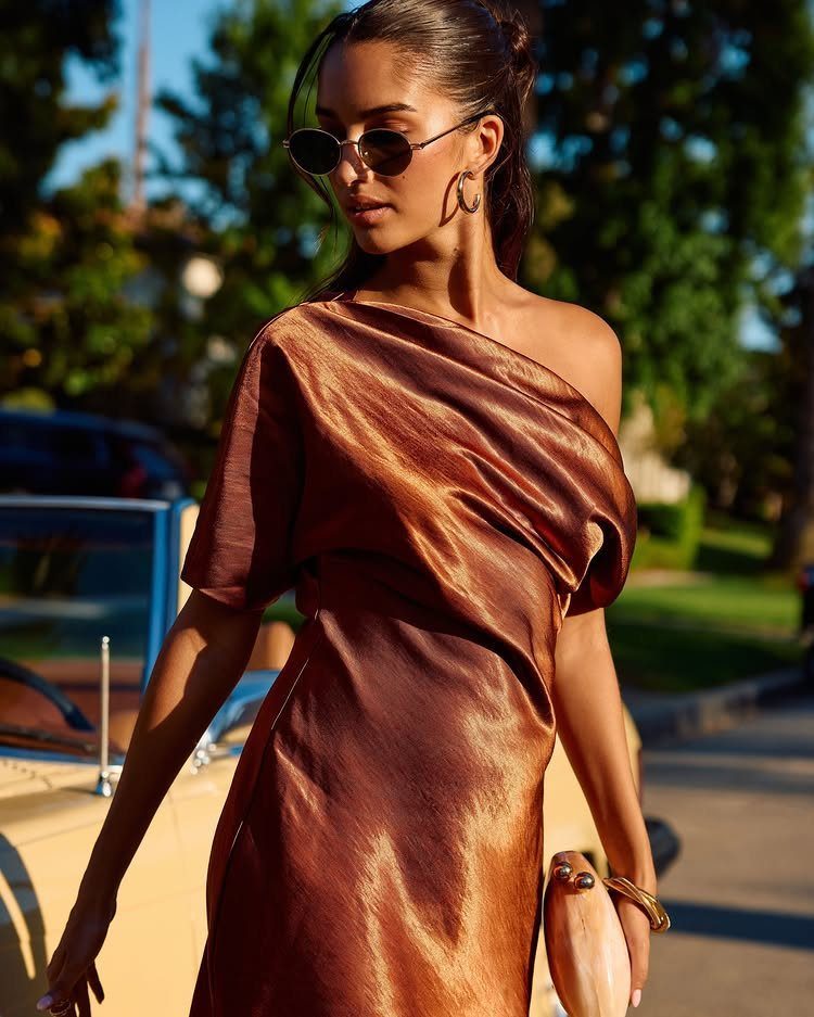 Woman in a bronze one-shoulder satin dress posing outdoors near a vintage car.