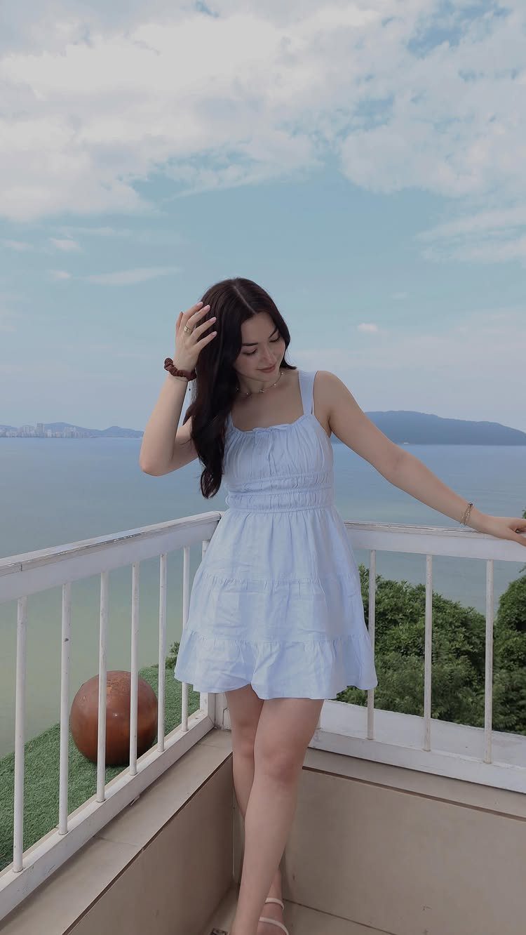 A woman in a light blue summer dress posing on a balcony overlooking the ocean.