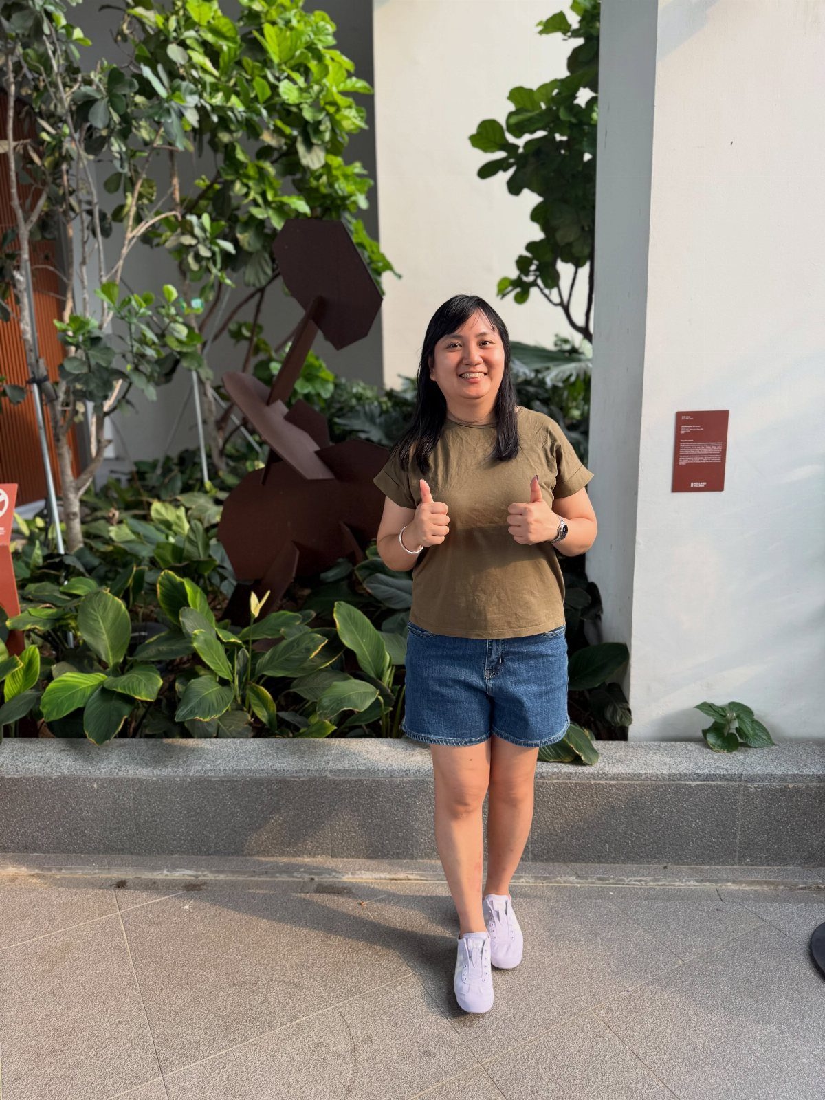 Woman in olive t-shirt and denim shorts giving two thumbs up outdoors.