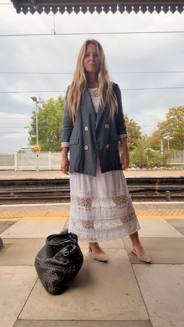 Woman in a white lace dress and striped blazer standing on a train platform.