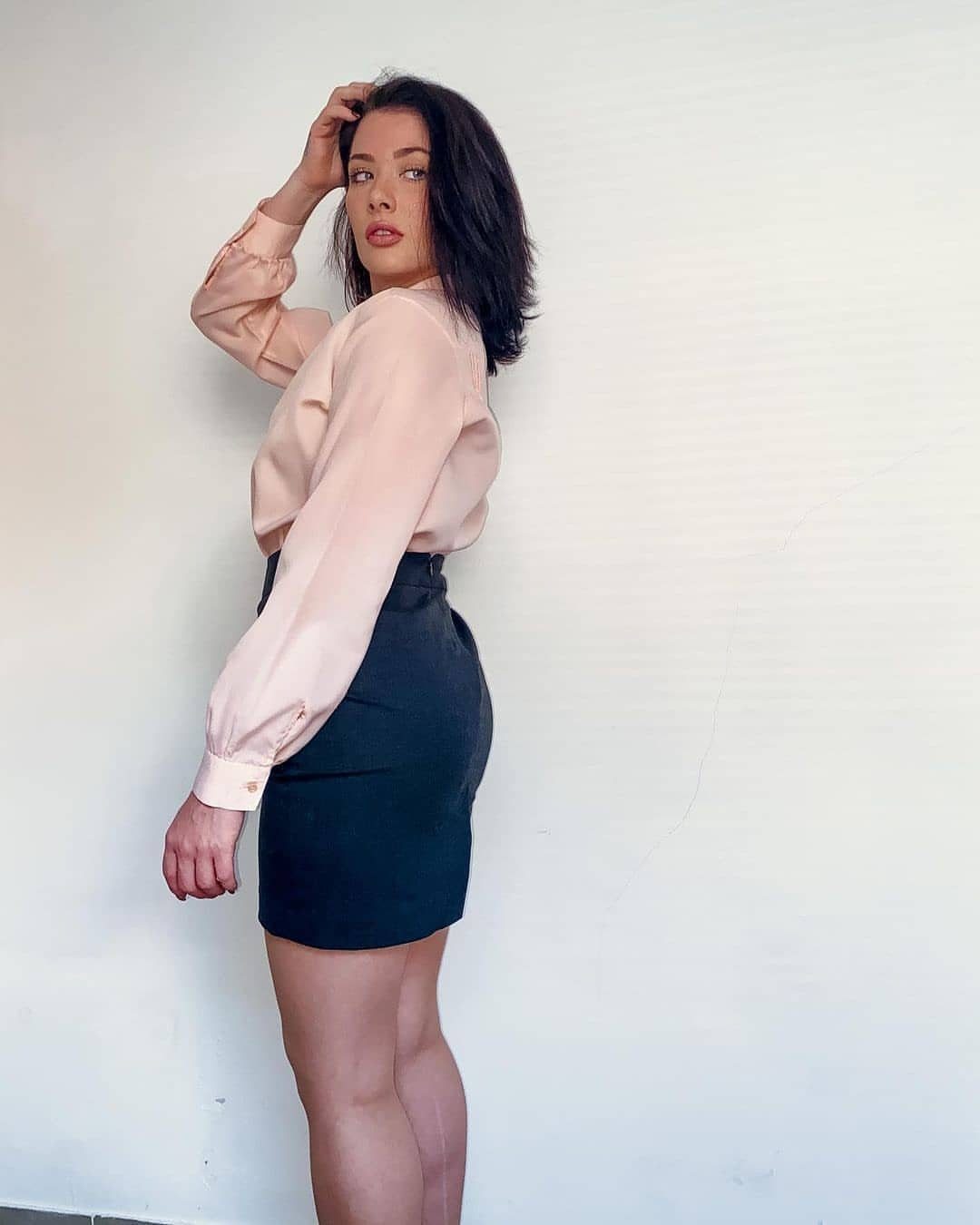 Woman in a pink button-up blouse and dark high-waisted mini skirt posing against a white wall.