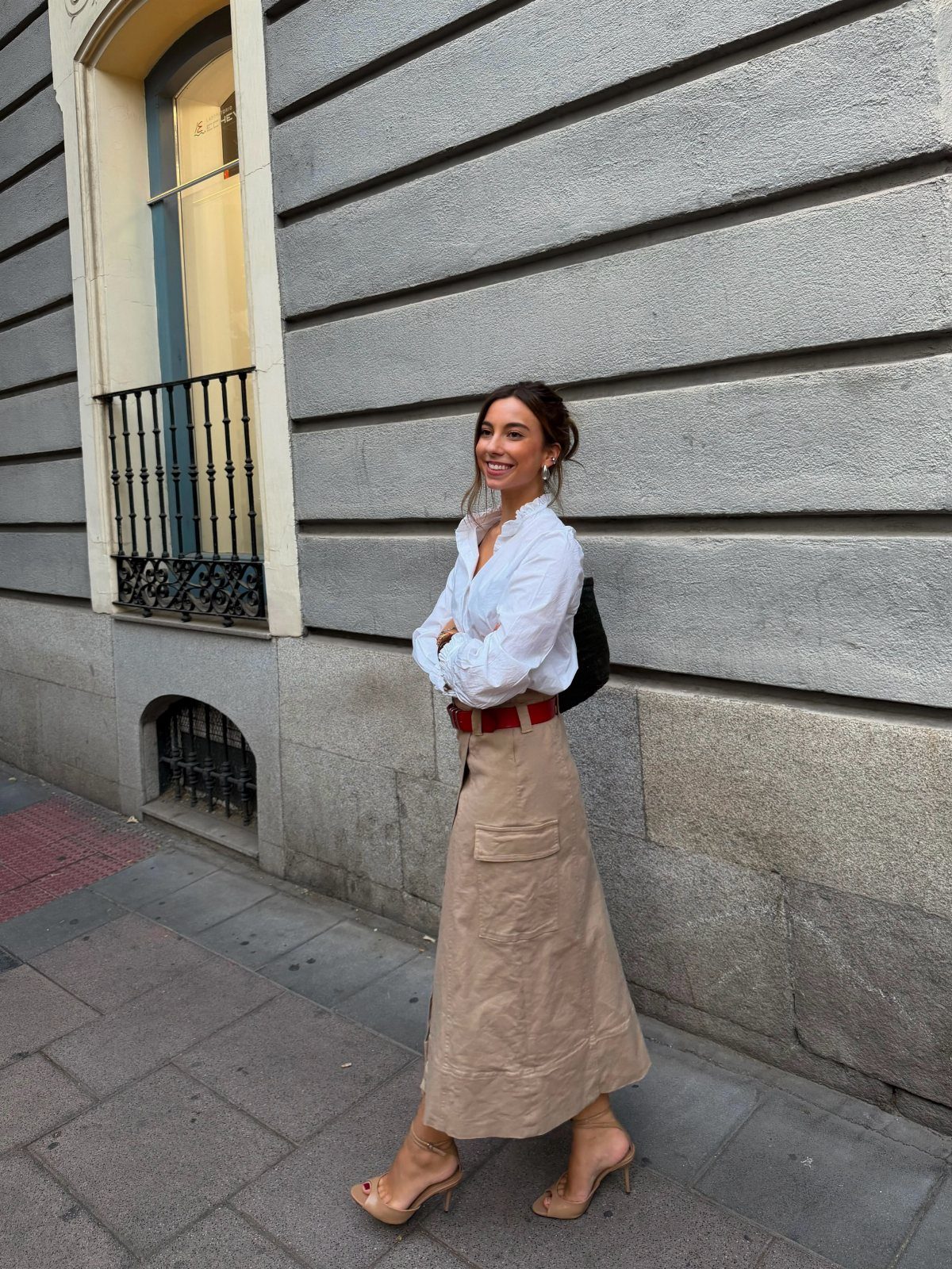 Woman walking in a white blouse, tan midi skirt, and red belt on a city street.