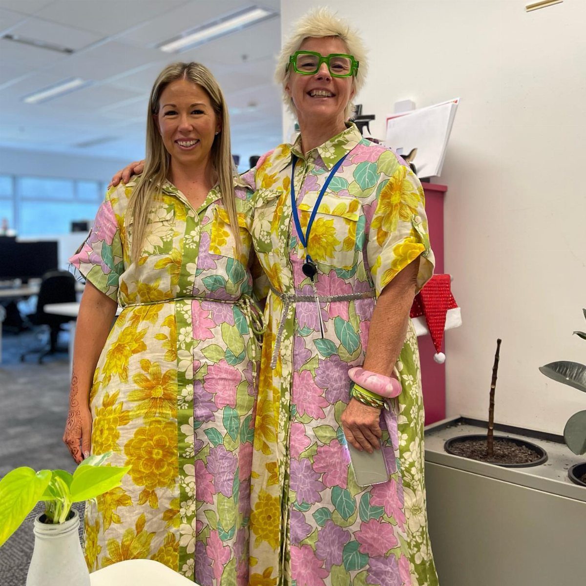 Two women smiling while wearing bright, floral-patterned shirtdresses in an office setting.