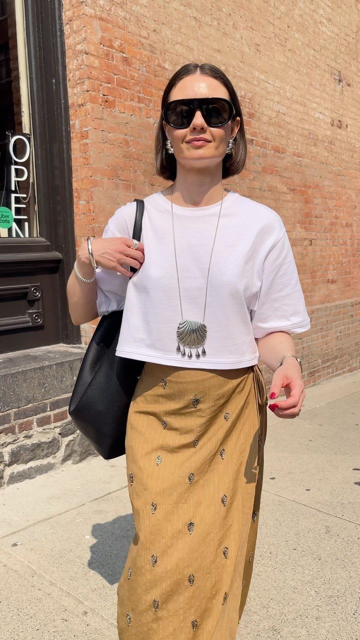 Woman in a white t-shirt and long mustard patterned skirt standing outdoors.