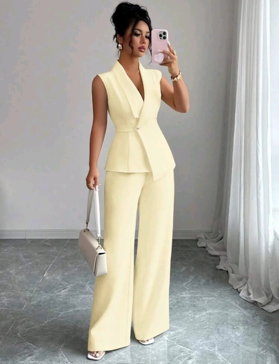 Woman in a pale yellow sleeveless vest and wide-leg trousers taking a mirror selfie.