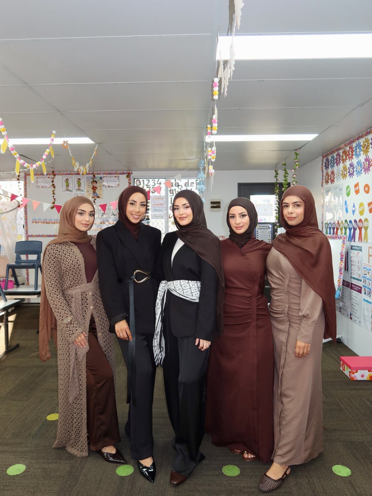 Five women in hijabs posing together in a classroom setting wearing modest outfits.