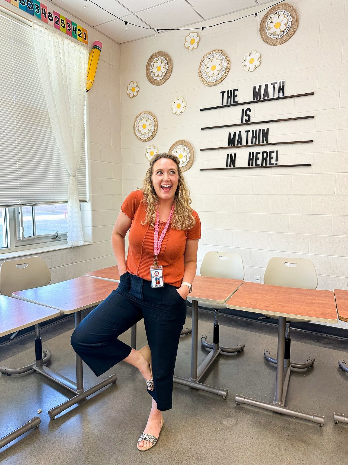 A smiling woman in an orange top and navy trousers posing in a classroom.