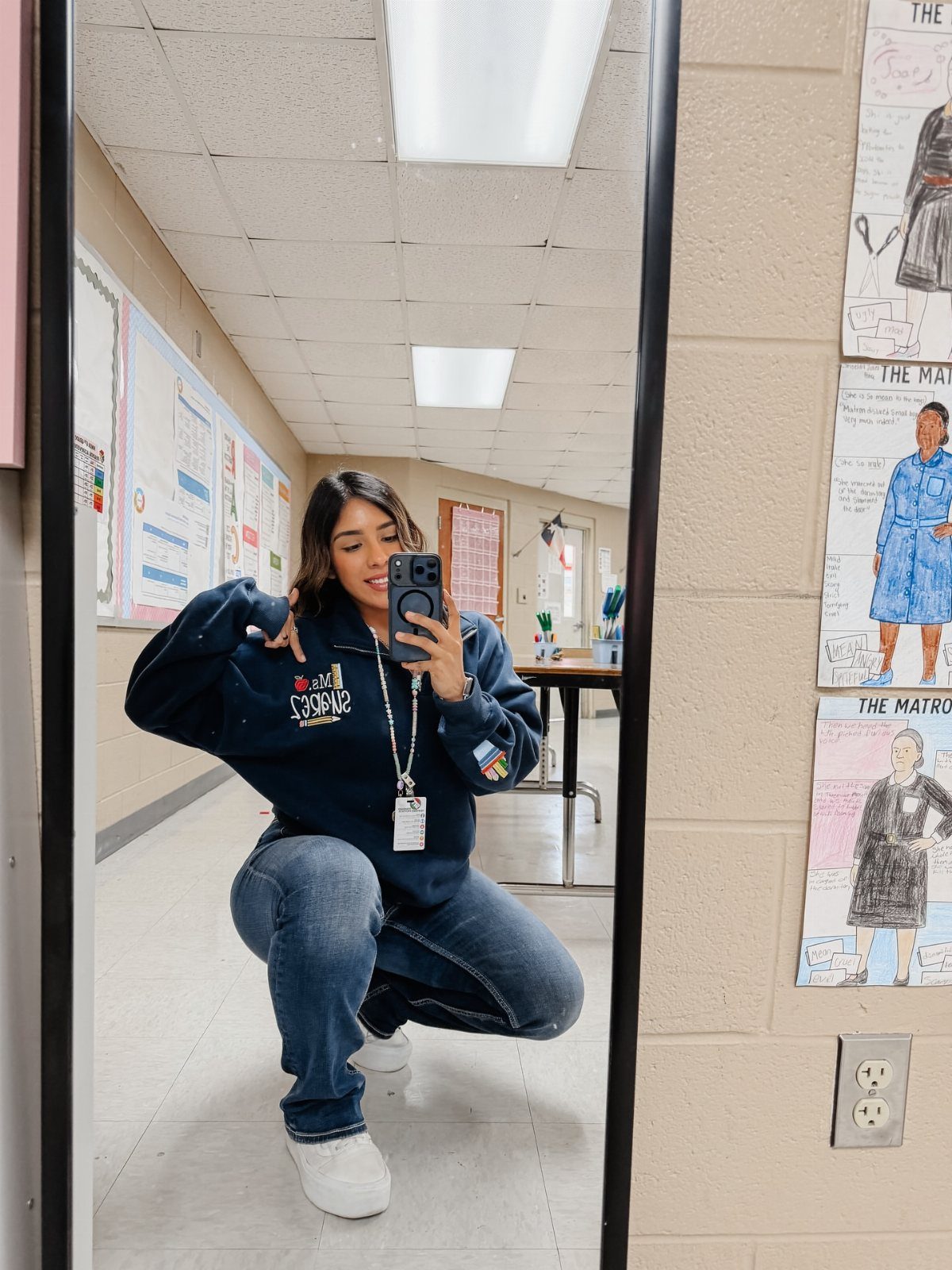 A woman in a navy graphic sweatshirt and blue jeans posing for a mirror selfie.