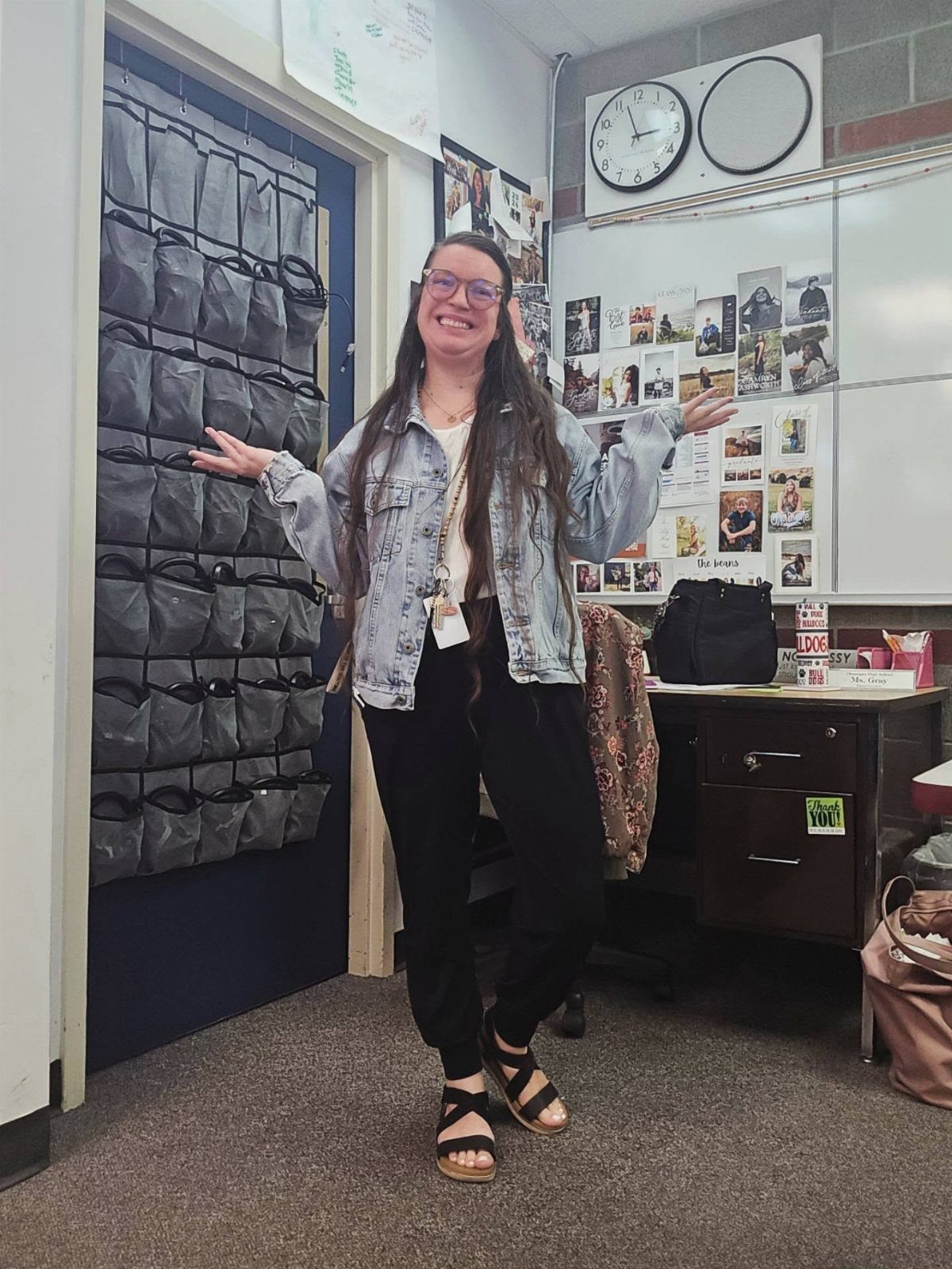 Smiling woman wearing a light blue denim jacket, white top, and black pants in a classroom.