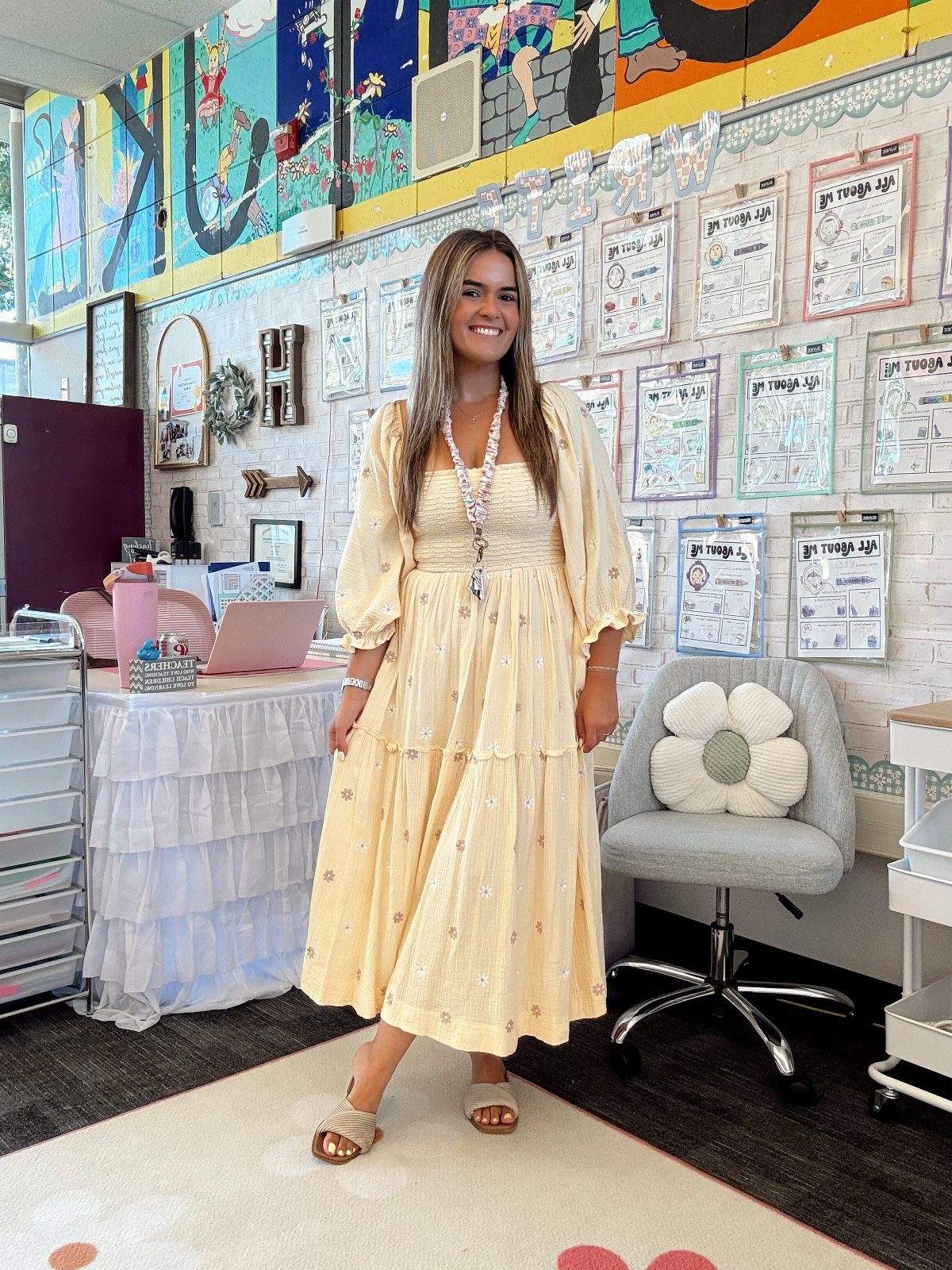 Smiling woman posing in a classroom wearing a pale yellow floral midi dress and sandals.