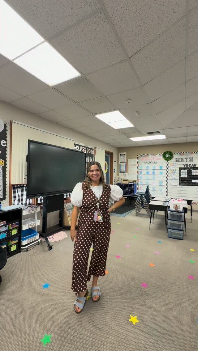 Woman in a classroom wearing a white blouse under a brown polka-dot jumpsuit.