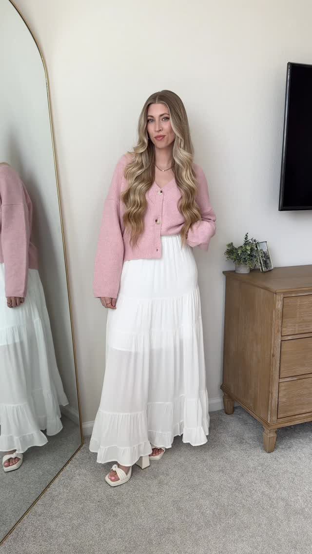Woman wearing a pink cardigan and a long white tiered maxi skirt standing in a room.