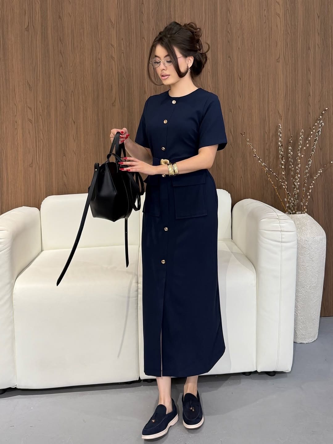 A woman standing in a navy blue midi dress while holding a black handbag.