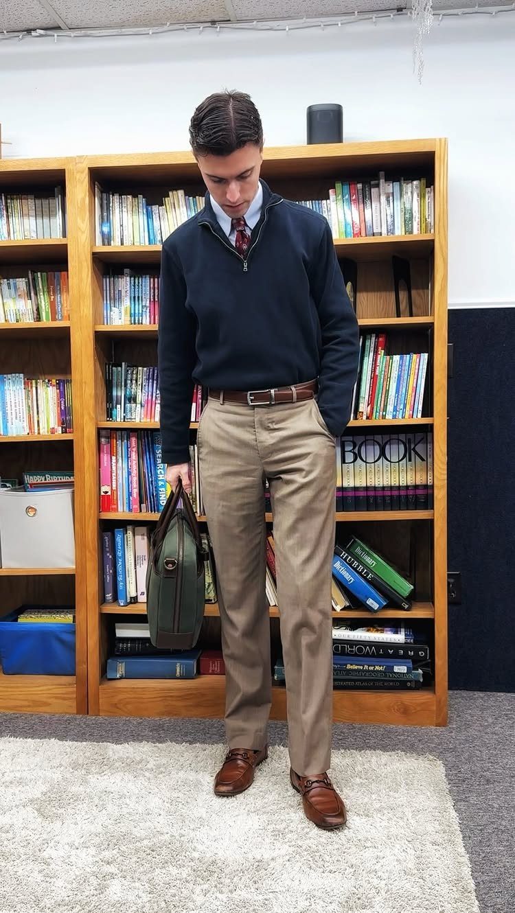 Man wearing a navy sweater, white shirt, red tie, and khaki trousers standing in a classroom.