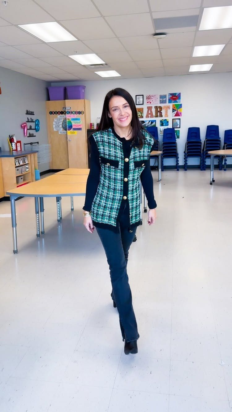 A smiling woman posing in a classroom wearing a plaid vest and dark flared pants.