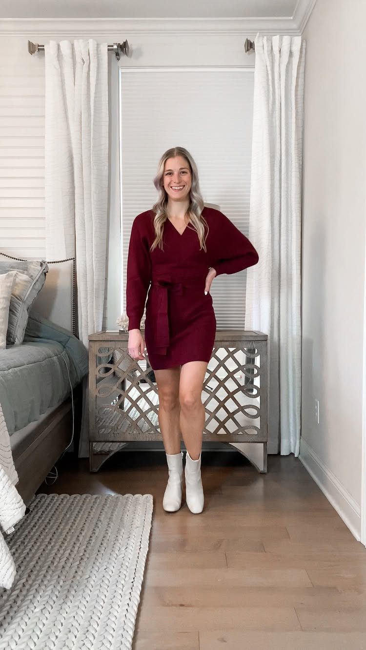 A woman in a maroon long-sleeved wrap dress and white boots standing in a bedroom.