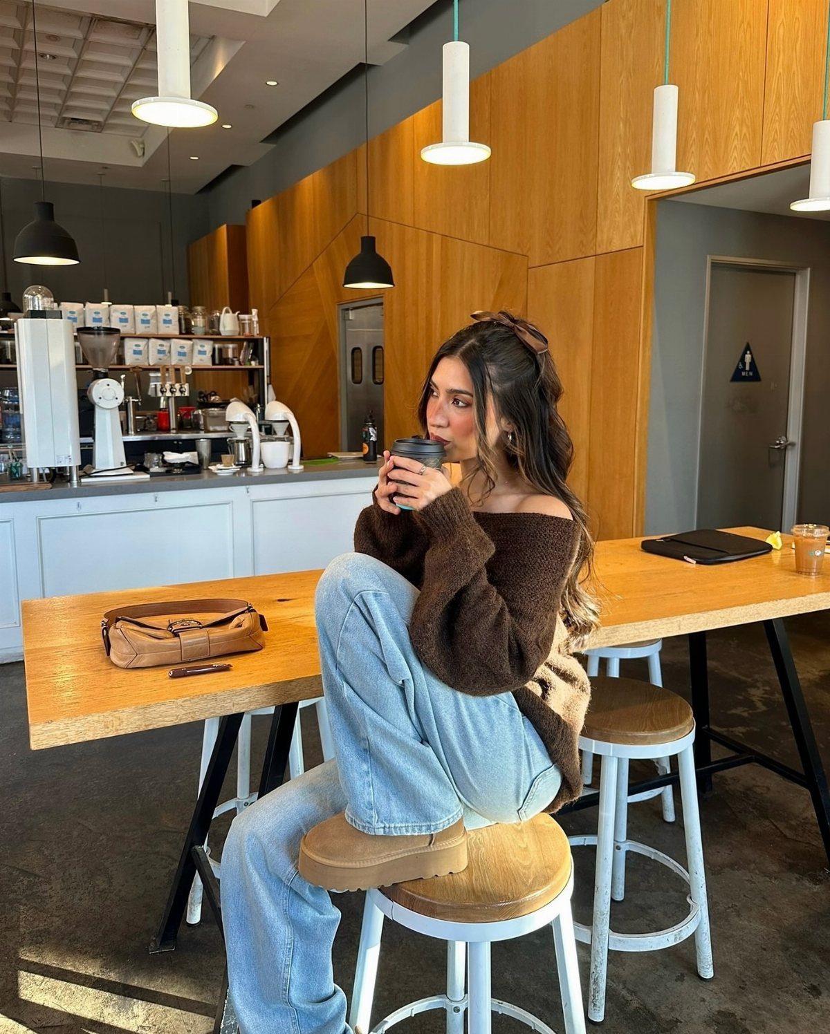 Woman in a brown sweater and light blue jeans sitting on a stool in a cafe.