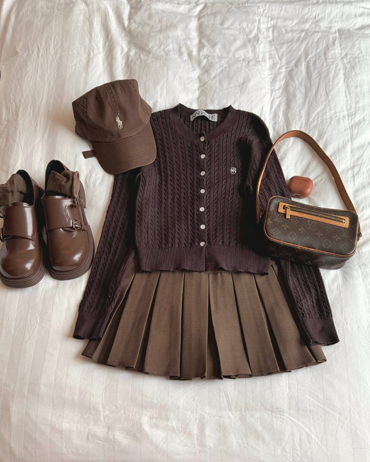 Dark brown cable-knit cardigan, pleated skirt, loafers, and accessories laid on white fabric.