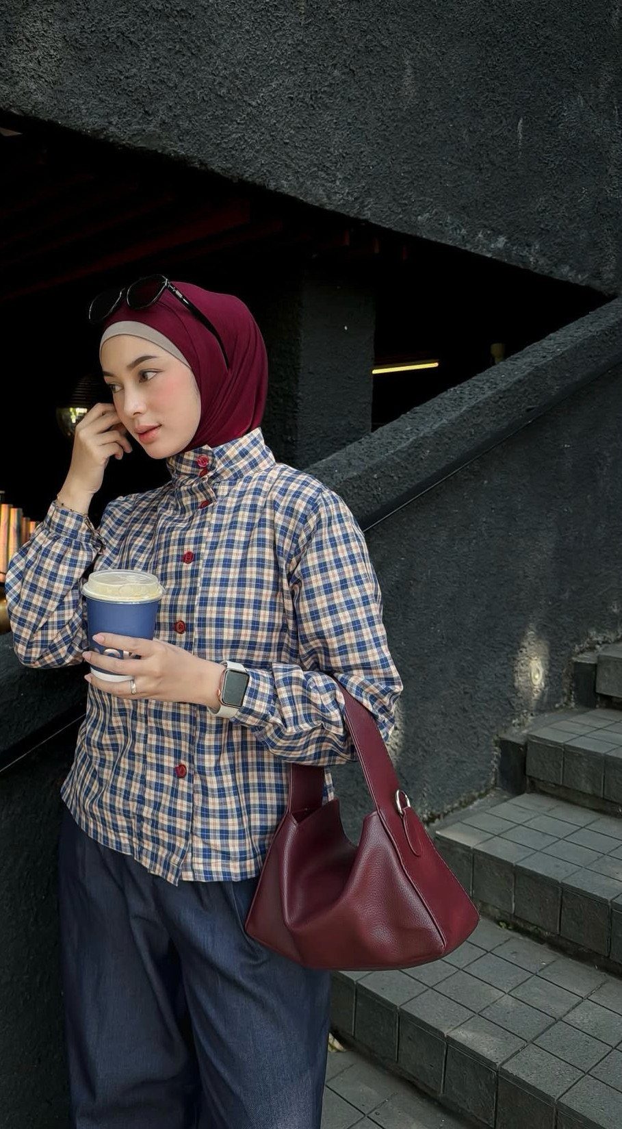 Woman in a blue plaid shirt and maroon hijab holding a coffee cup.