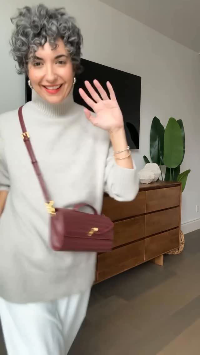 Woman with curly grey hair wearing a white turtleneck sweater and waving.