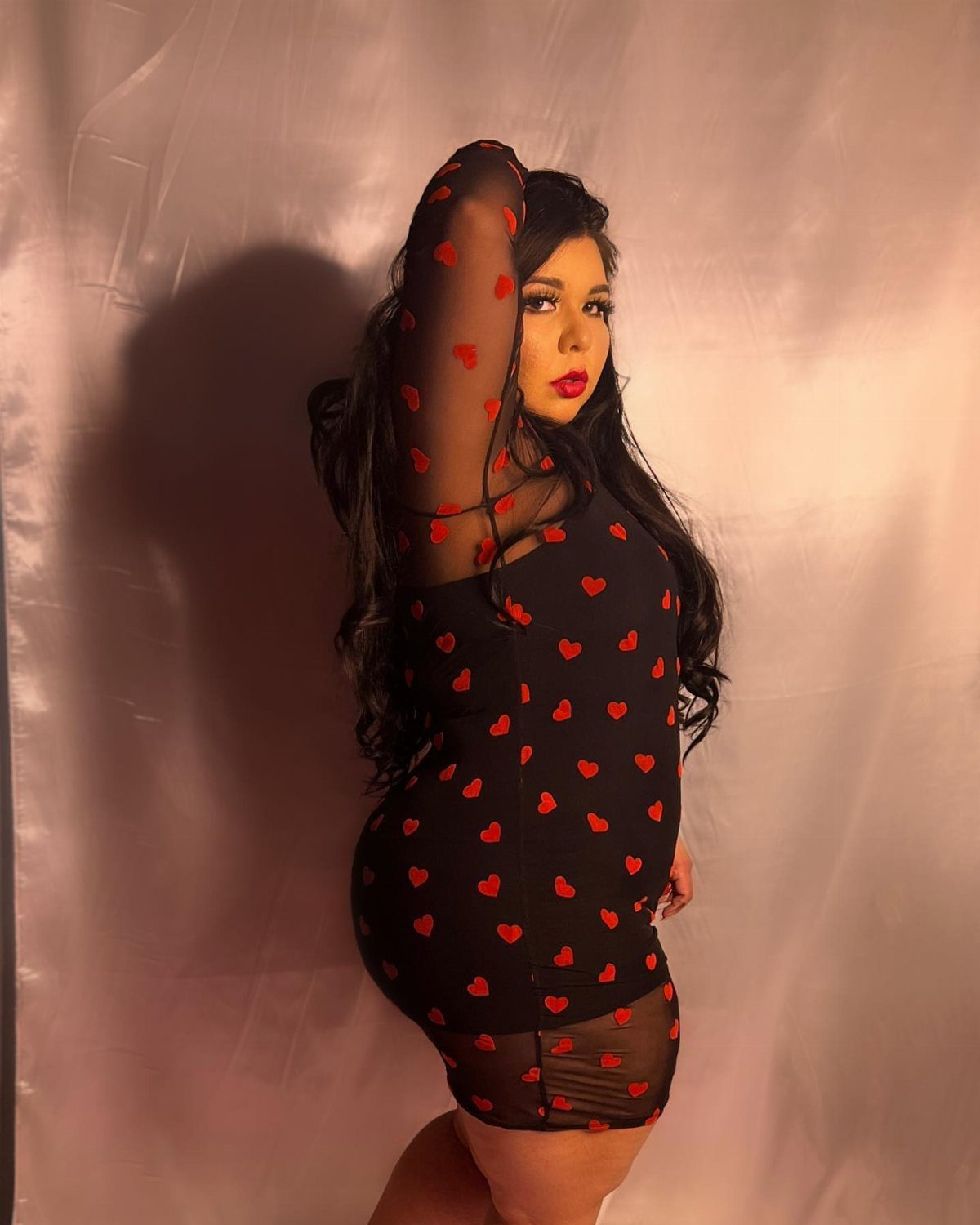 Woman posing in a black mini dress with a red heart print and sheer sleeves.