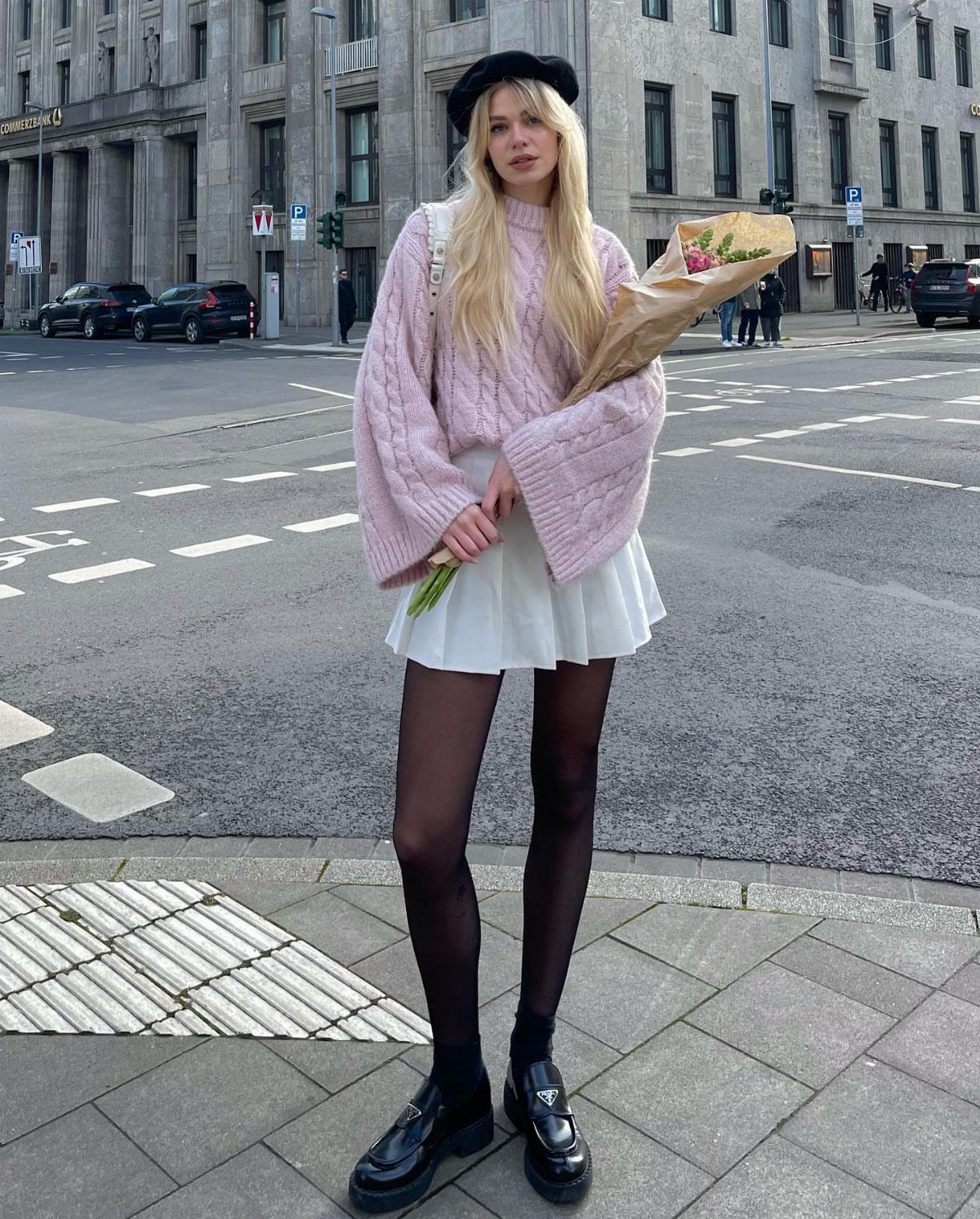 Blonde woman wearing a pink oversized sweater and white mini skirt holding flowers.