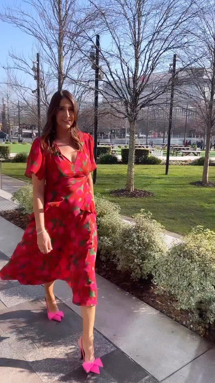 A woman in a red floral midi dress and pink heels standing on a sidewalk.