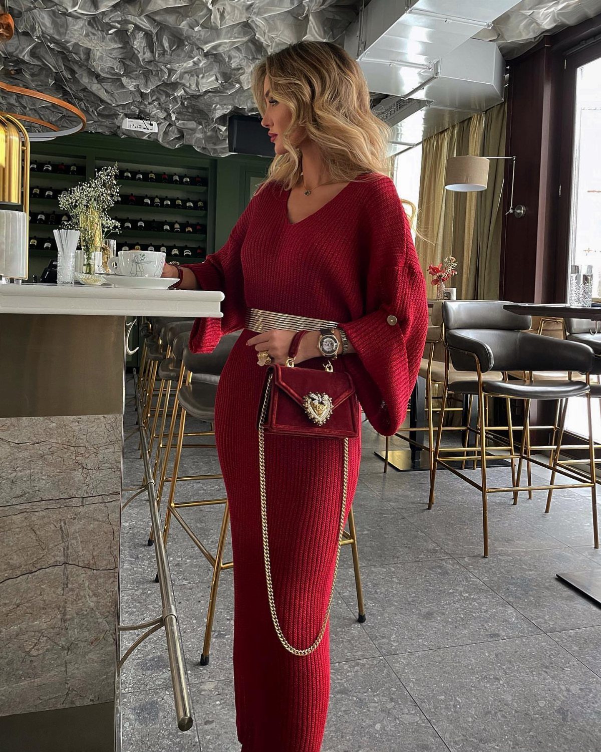 A blonde woman wearing a long burgundy ribbed knit maxi dress and holding a red heart-shaped handbag.