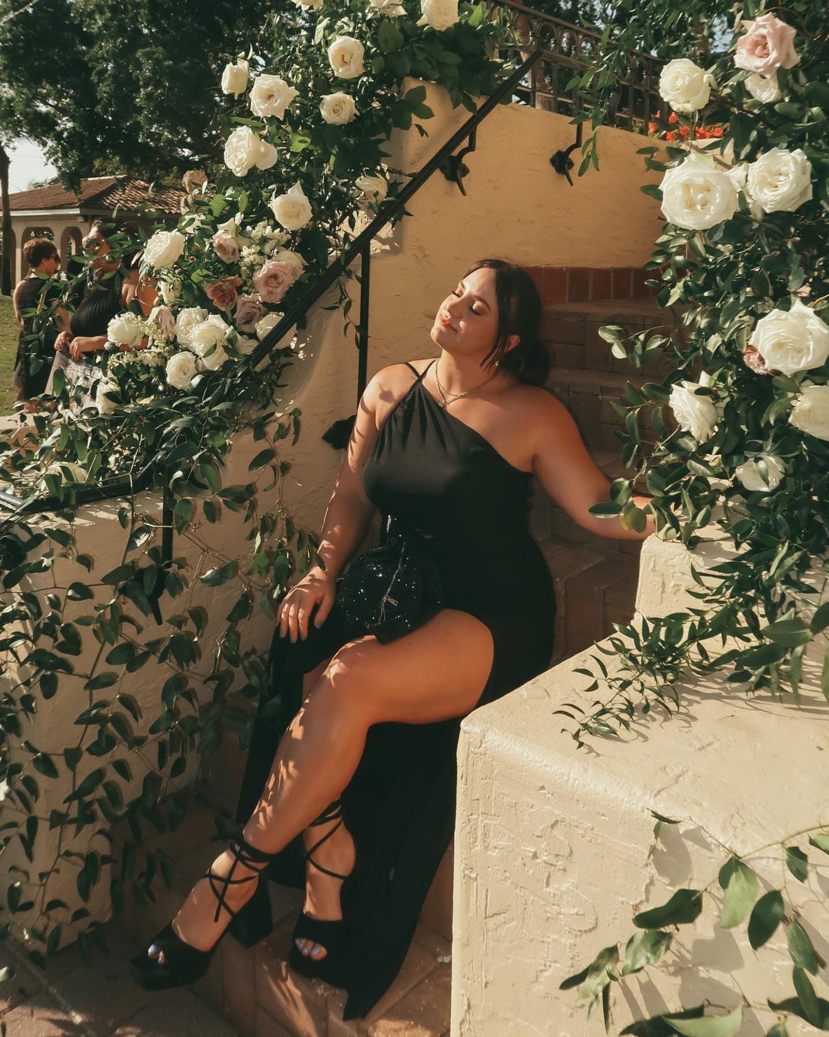 A woman in a black one-shoulder formal gown sitting on stone stairs surrounded by white roses.