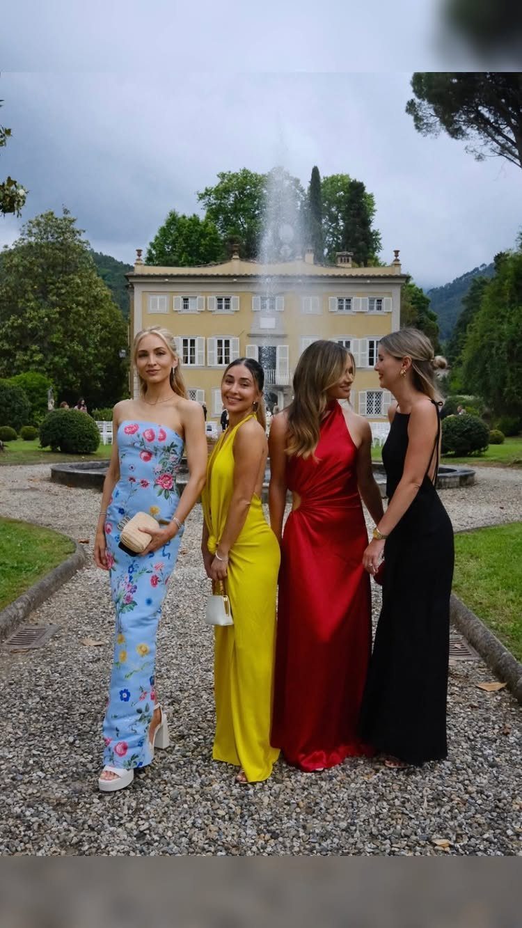 Four women in long evening gowns posing in a garden in front of a large villa.