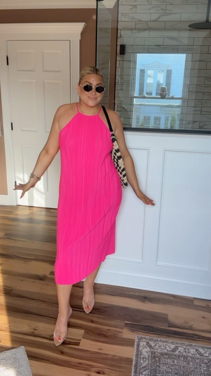 Woman in a bright pink halter midi dress and clear heels posing indoors.
