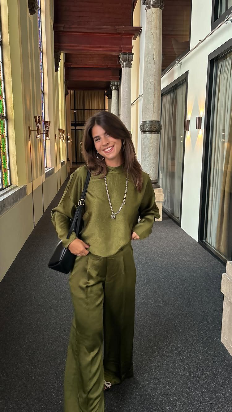 A woman smiling in an olive green silk top and wide-leg trousers standing in a hallway.
