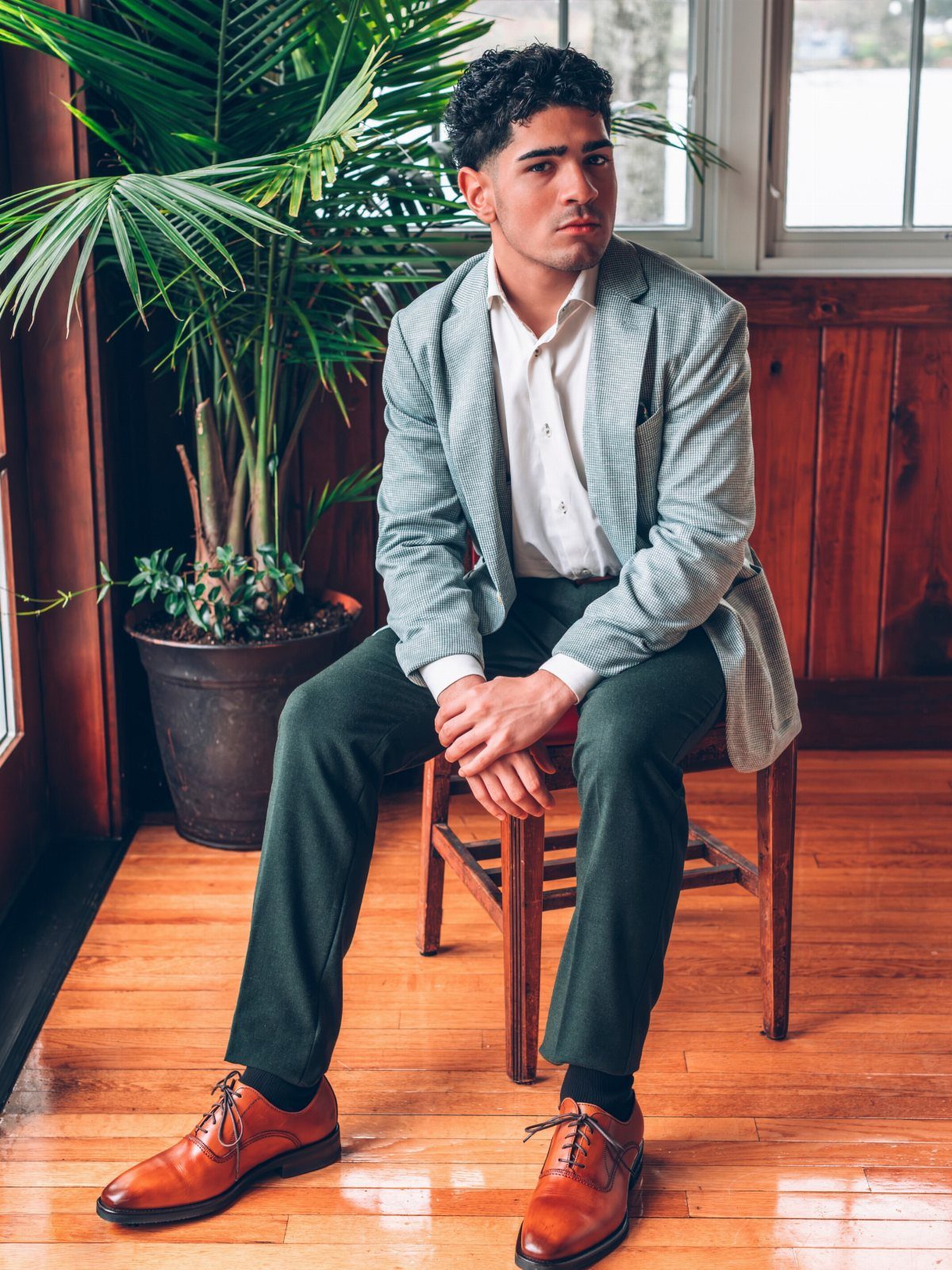 A young man posing in a light grey blazer, white shirt, green trousers, and brown shoes.