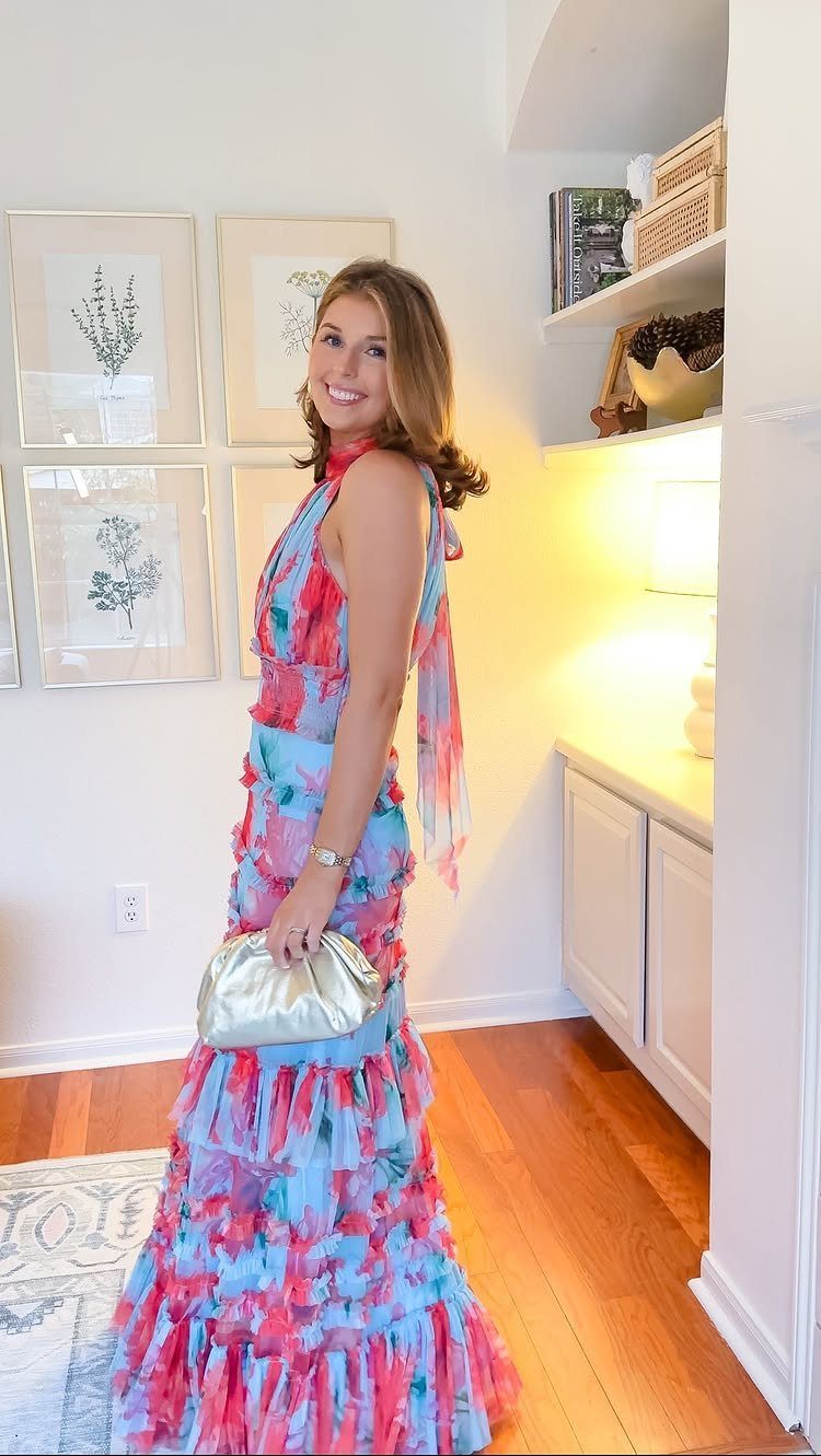 A woman in a colorful pink and blue ruffled maxi dress posing with a gold clutch.