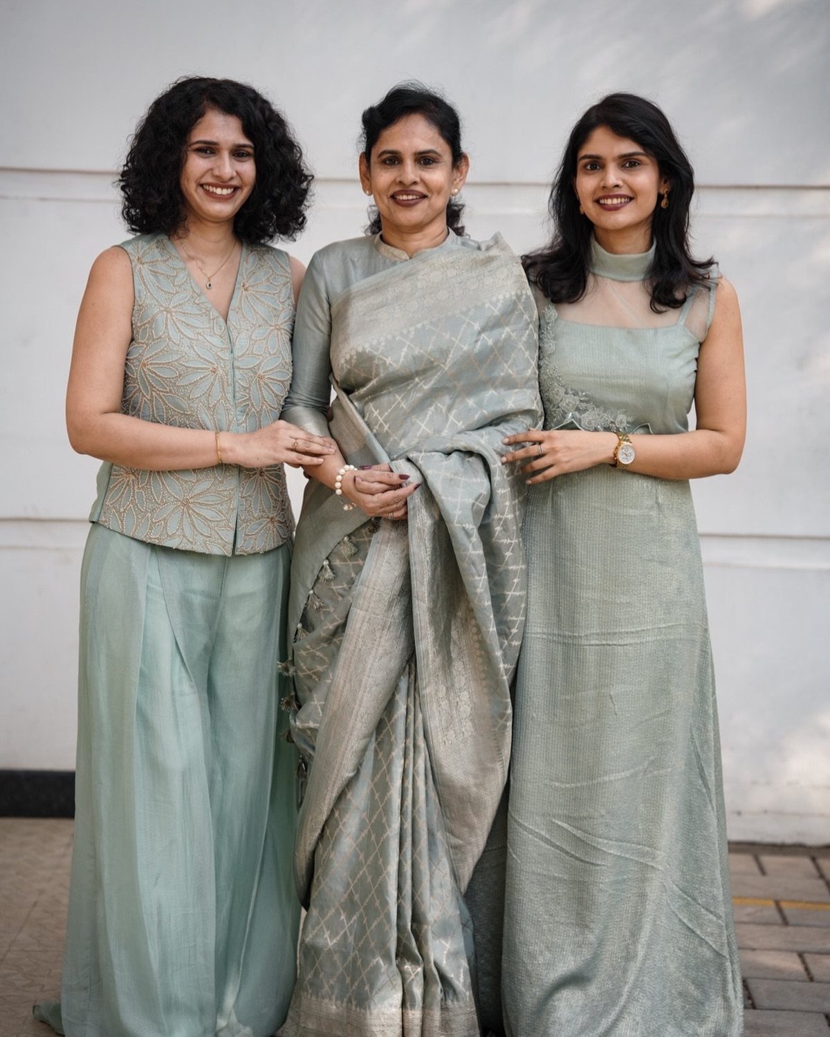 Three women smiling while wearing elegant sage green traditional Indian outfits against a white wall.
