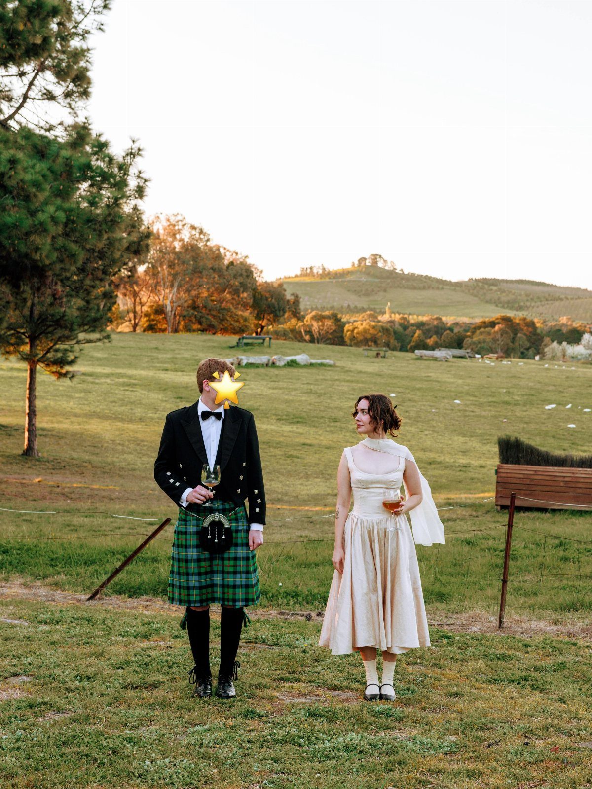 A man in a formal kilt and a woman in a cream midi dress standing in a field.