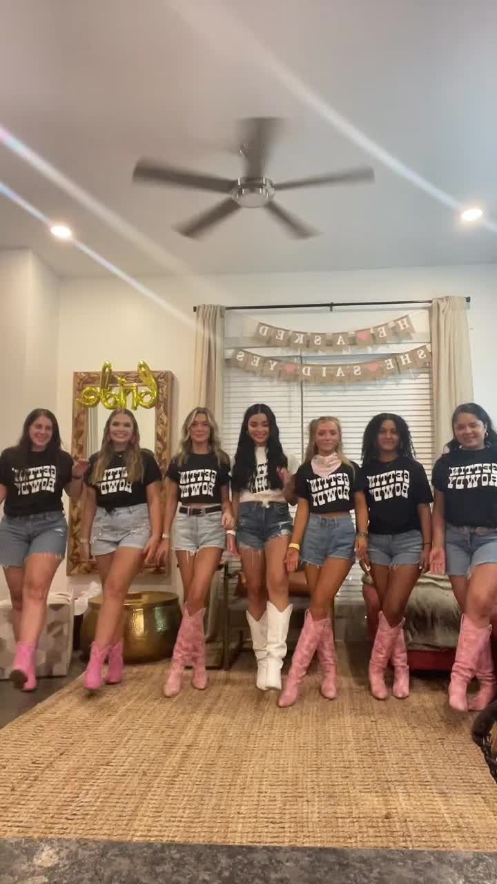 Seven women posing in matching black t-shirts, denim shorts, and cowboy boots in a living room.