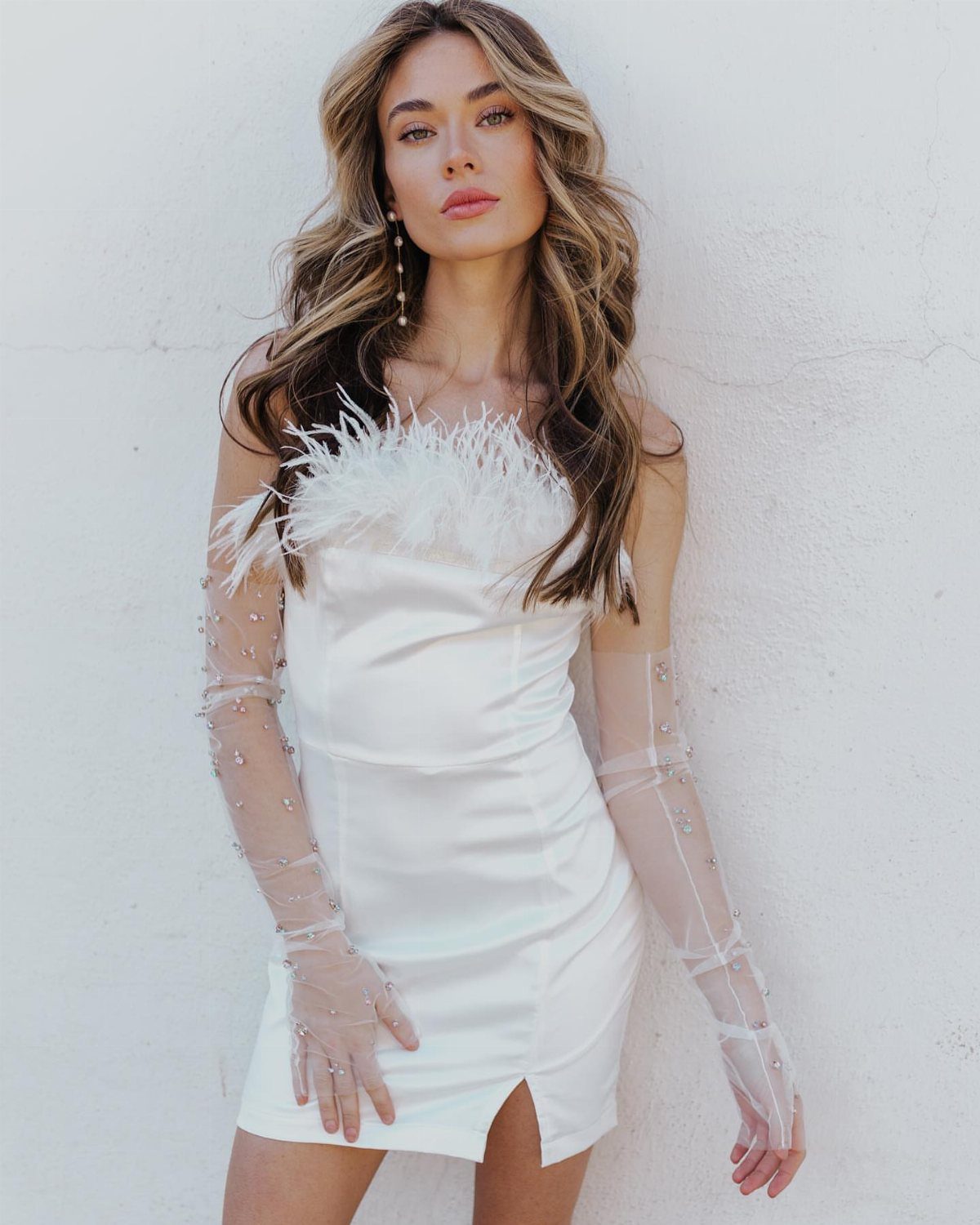 Woman in a white satin mini dress with feather trim and sheer pearl-embellished gloves posing against a white wall.