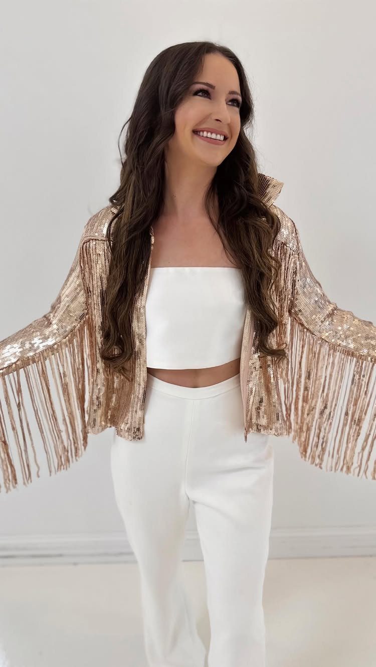 Smiling woman wearing a white crop top, white trousers, and a rose gold sequined fringe jacket.