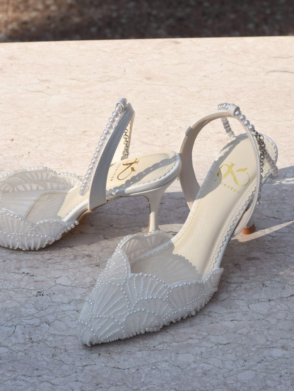 White pearl-encrusted kitten heel sandals resting on a light stone surface outdoors.