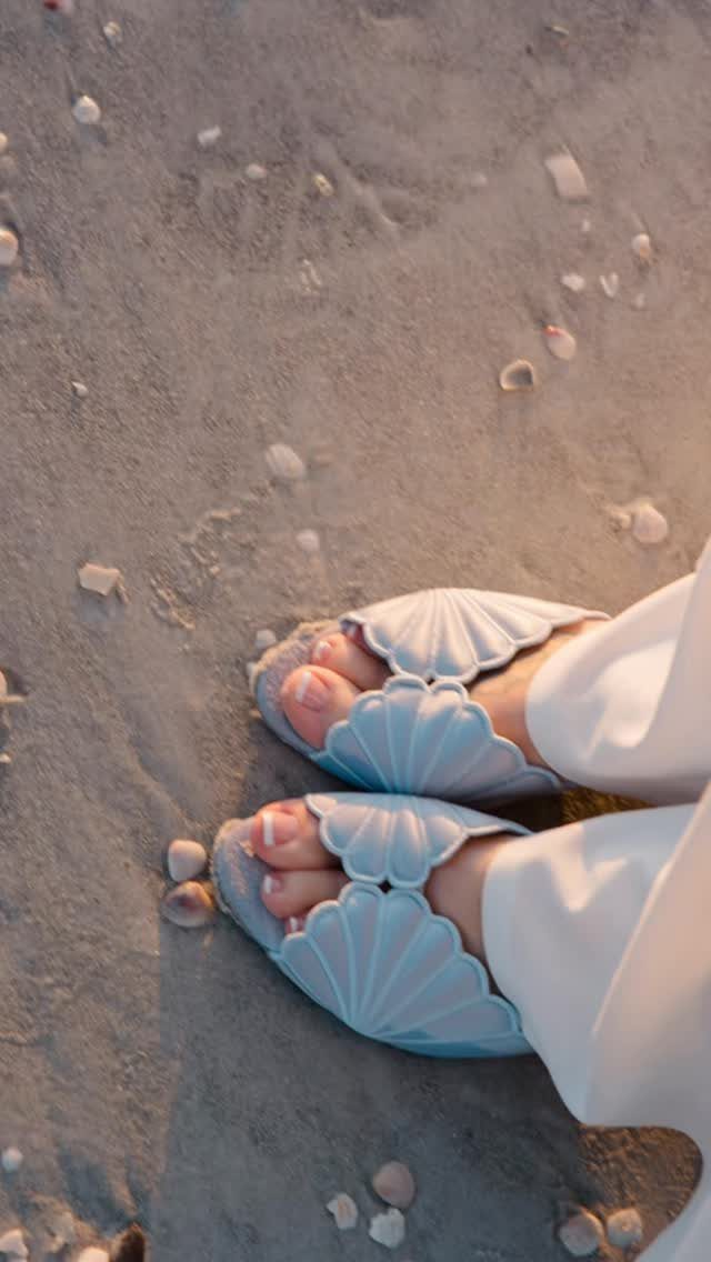 Light blue shell-shaped sandals worn by a person walking on a sandy beach with seashells.