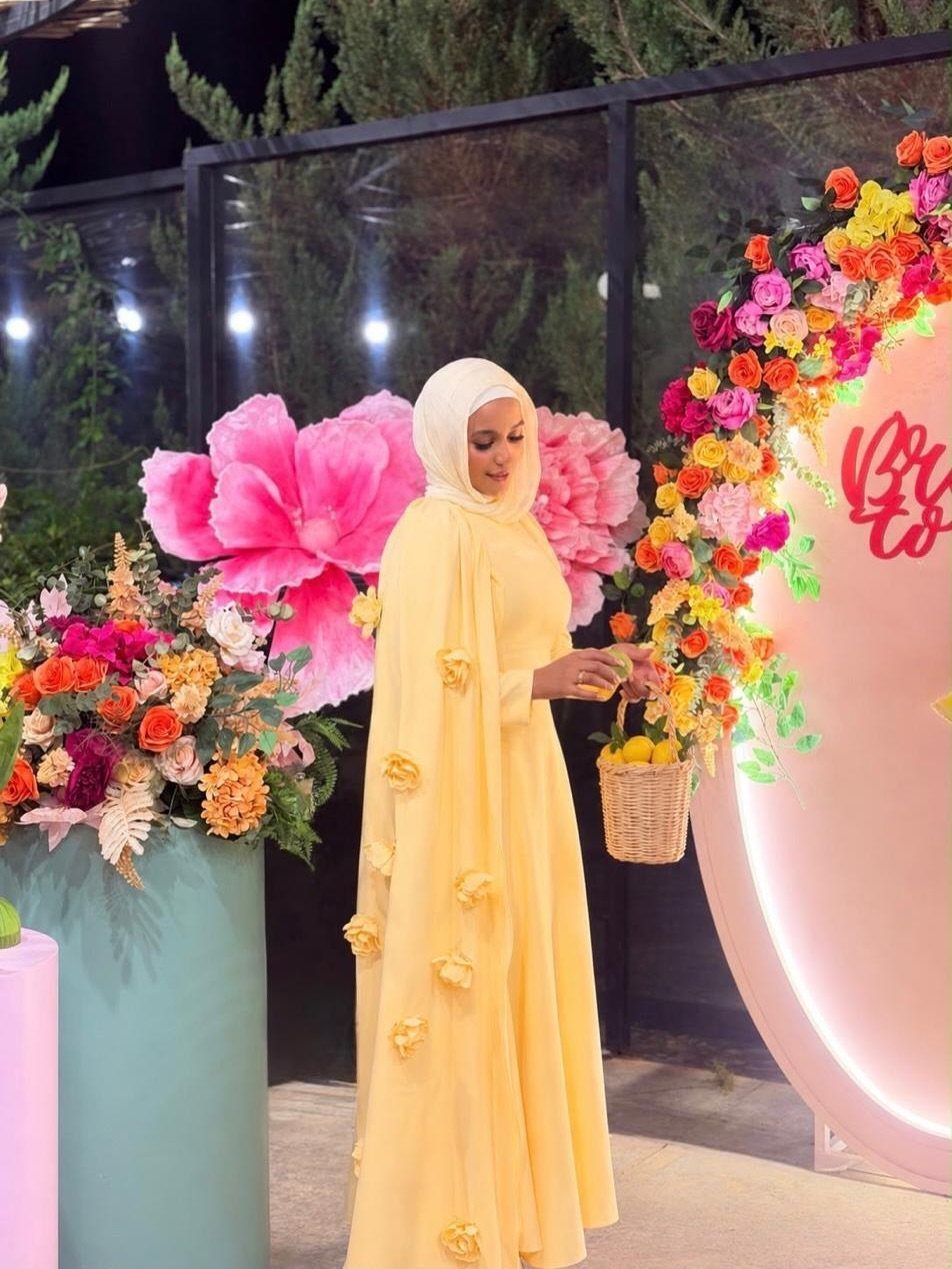Woman in a long yellow flowing gown and white hijab standing by large pink flowers.