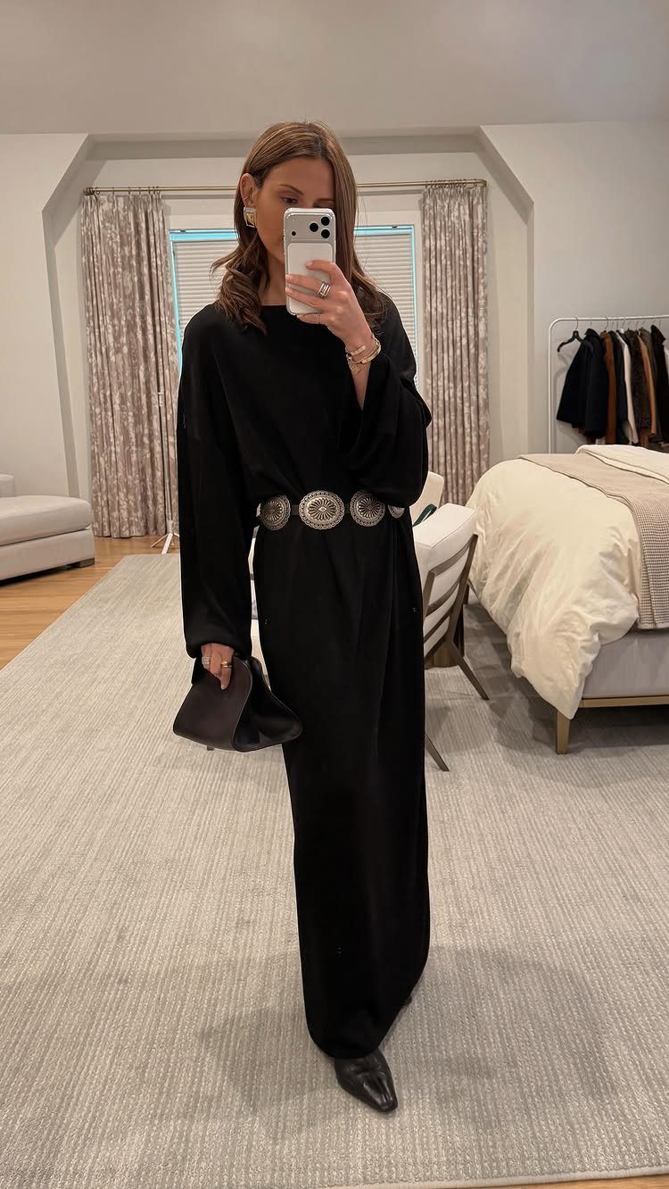 Woman in a long black maxi dress with a silver belt posing for a mirror selfie.