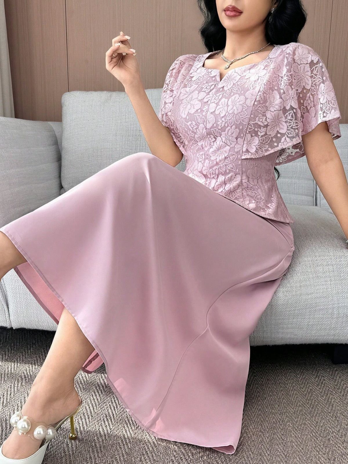 Woman in a pink lace top and satin midi skirt sitting on a grey sofa.