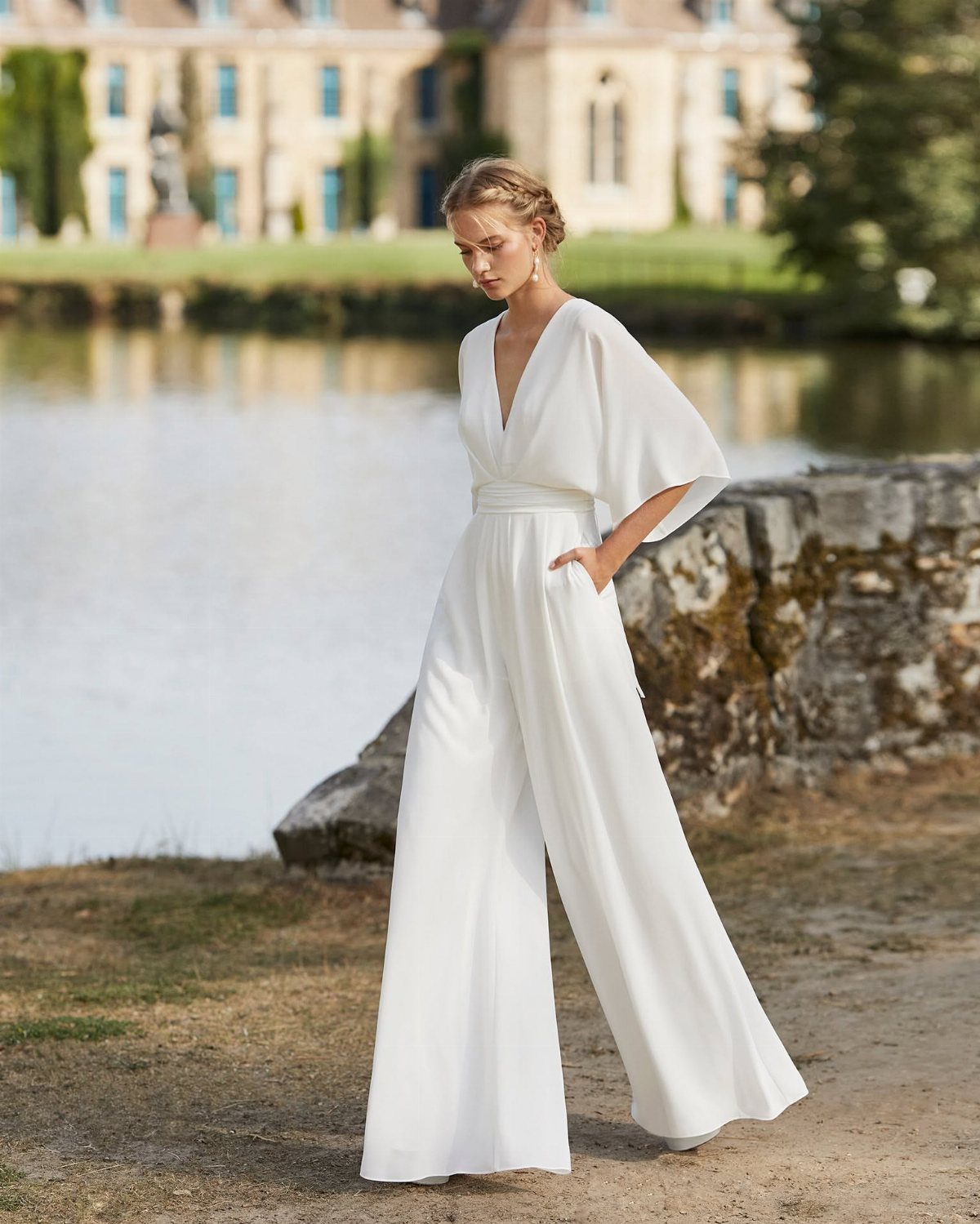 Woman in a white wide-leg jumpsuit standing by a lake near a large building.